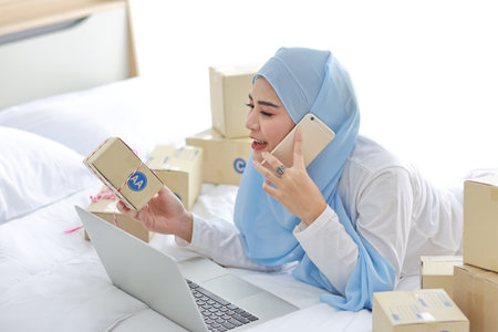 Beautiful and young asian woman in muslim sleepwear with attractive look, lies on bed with computer and online package box delivery. Startup small business SME freelance girl working with mobile phoneの写真素材