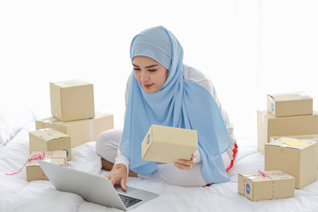 Beautiful and young asian woman in muslim sleepwear with attractive look, sitting on bed with computer and online package box delivery. Startup small business SME freelance girl work with mobile phoneの写真素材