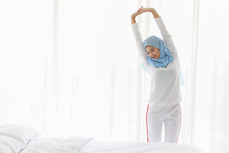 Beautiful asian woman wearing white muslim sleepwear, stretching her arms after getting up in the morning at sunrise. Cute young girl with blue hijab standing and relaxing with happy and smiling face.の写真素材