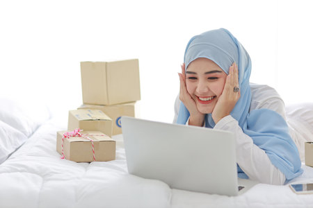Beautiful and young asian woman in muslim sleepwear with attractive look, lies on bed with computer, mobile phone and online package box delivery. Smart girl with hijab receive good news and surprise.の写真素材