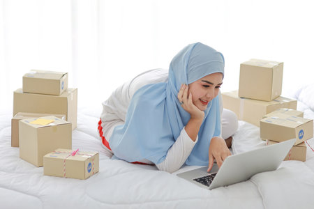 Beautiful and young asian woman in muslim sleepwear with attractive look, lies on bed with computer and online package box delivery. Startup small business SME freelance girl working with mobile phoneの写真素材