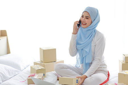 Beautiful and young asian woman in muslim sleepwear with attractive look, sitting on bed with computer and online package box delivery. Startup small business SME freelance girl work with mobile phoneの写真素材