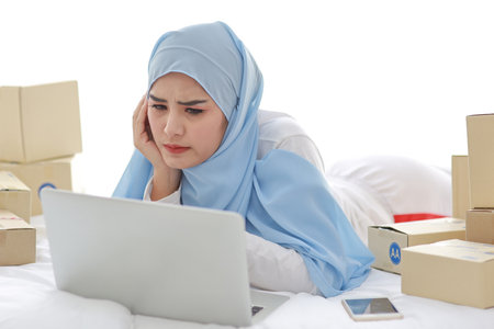 Beautiful and young asian woman in muslim sleepwear with headache emotion lies on bed with computer, mobile phone and online package box delivery. Smart girl with hijab receive bad news and tiredの写真素材