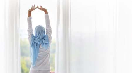Rear view beautiful asian woman wearing white muslim sleepwear, stretching her arms after getting up in the morning at sunrise. Cute young girl with blue hijab standing and relaxing while looking awayの写真素材
