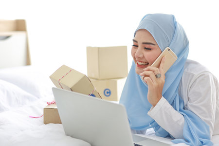 Beautiful and young asian woman in muslim sleepwear with attractive look, lies on bed with computer and online package box delivery. Startup small business SME freelance girl working with mobile phoneの写真素材