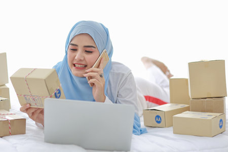 Beautiful and young asian woman in muslim sleepwear with attractive look, lies on bed with computer and online package box delivery. Startup small business SME freelance girl working with mobile phoneの写真素材
