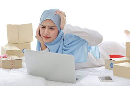 Beautiful and young asian woman in muslim sleepwear with headache emotion lies on bed with computer, mobile phone and online package box delivery. Smart girl with hijab receive bad news and tiredの写真素材