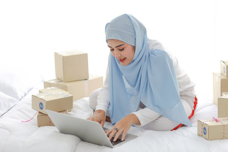 Beautiful and young asian woman in muslim sleepwear with attractive look, sitting on bed with computer and online package box delivery. Startup small business SME freelance girl work with mobile phoneの写真素材