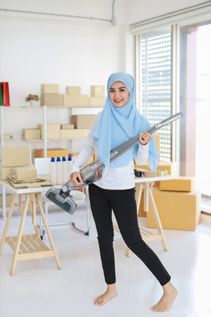Active young beautiful asian housewife woman cleaning with vacuum cleaning floor after working SME with E-commerce Package box background. Cleaning girl in muslim black leggings vacuuming her room.の写真素材