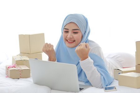 Beautiful and young asian woman in muslim sleepwear with attractive look, lies on bed with computer, mobile phone and online package box delivery. Smart girl with hijab receive good news and surprise.の写真素材