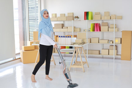Active young beautiful asian housewife woman cleaning with vacuum cleaning floor after working SME with E-commerce Package box background. Cleaning girl in muslim black leggings vacuuming her room.の写真素材