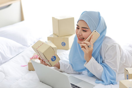 Beautiful and young asian woman in muslim sleepwear with attractive look, lies on bed with computer and online package box delivery. Startup small business SME freelance girl working with mobile phoneの写真素材