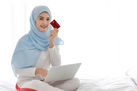 Young asian woman sitting on bed and holding credit card with computer in bedroom. Beautiful girl in muslim sleepwear with hijab purchasing or online payment. Lifestyle and technology conceptの写真素材
