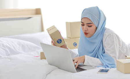 Beautiful and young asian woman in muslim sleepwear with attractive look, lies on bed with computer and online package box delivery. Startup small business SME freelance girl working with mobile phoneの写真素材