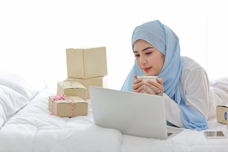 Beautiful and young asian woman in muslim sleepwear with attractive look, lies on bed with computer and online package box delivery. Startup small business SME freelance girl working with coffee cupの写真素材