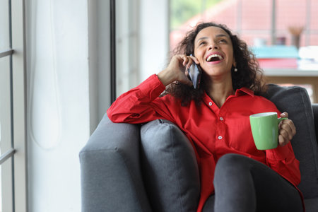 Happy and smiling latin woman sitting on sofa and using internet on computer with smart mobile phone in living room. Lifestyle with technologyの写真素材