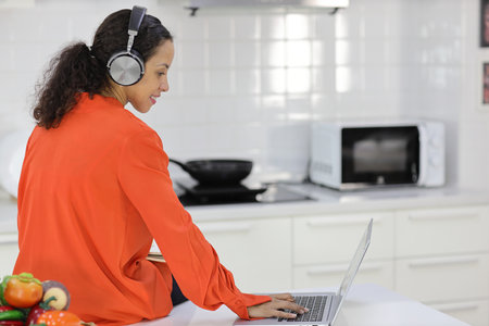 Happy latin woman learn cooking food while checking recipe from internet with computer in kitchen. Young female with headphone prepare delicious meal and listen music. Healthy and lifestyle conceptの写真素材
