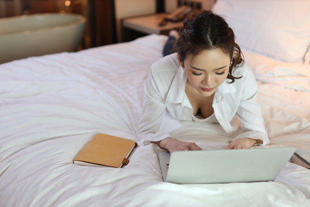 Active asian woman in casual lying on bed and working on computer with notebook in hotel, beauty businesswoman with bedroom background. Lifestyle technology conceptの写真素材