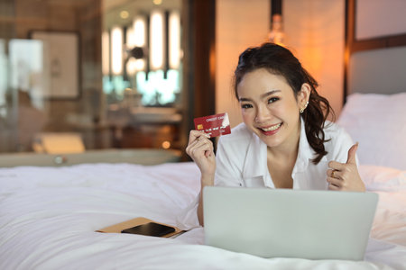 Happy asian woman lying on bed and holding credit card while using computer and show thumb up in hotel for online shopping payment, beauty businesswoman with bedroom. Lifestyle technology conceptの写真素材