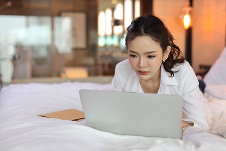 Active asian woman in casual lying on bed and working on computer with notebook in hotel, beauty businesswoman with bedroom background. Lifestyle technology conceptの写真素材