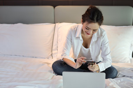 Active and happy asian woman in casual sitting on bed and working smart mobile phone and computer in hotel, beauty businesswoman with bedroom background. Lifestyle technology conceptの写真素材
