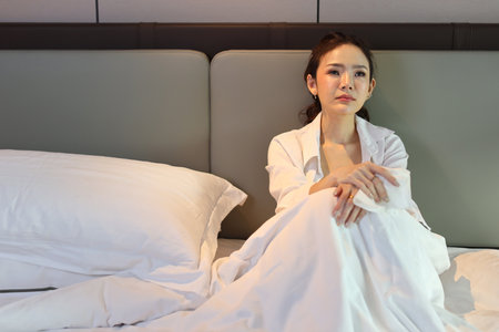 Young depressed asian woman sitting and looking at the ceiling while crying on bed in hotel bedroom with low light environment. Sad girl, unhappy and loneliness concept.の写真素材