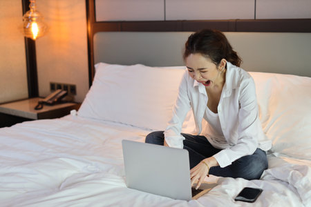 Active asian woman in casual sitting on bed and working smart mobile phone and computer while yawning in hotel, beauty businesswoman with bedroom background. Lifestyle technology conceptの写真素材