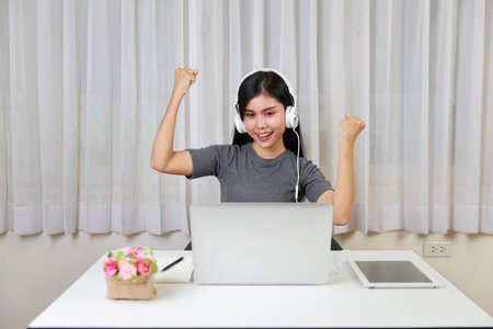 Young smart and active asian female student sitting at table with headset, using computer and taking note for studying online with exciting emotion (study online concept)の写真素材