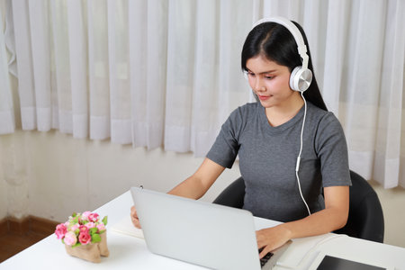 Young smart and active asian female student sitting at table with headset, using computer and taking note for studying online with exciting emotion (study online concept)の写真素材