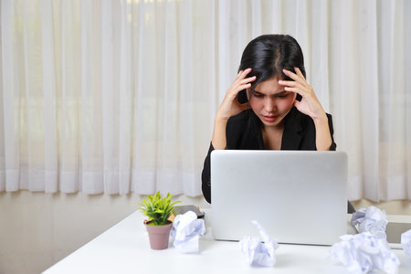 Mid adult businesswoman touching head on table after bad news business failure or get fired and feeling discouraged, distraught and hopeless on blue background with computer and crumpled paperの写真素材