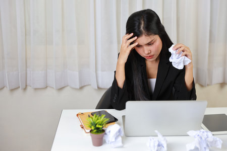 Mid adult businesswoman feeling upset  after bad news business failure or get fired and feeling discouraged, distraught and hopeless on blue background with computer and crumpled paperの写真素材