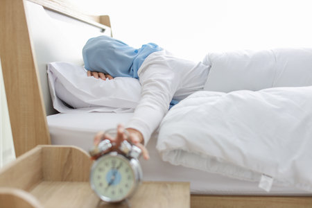 Oversleeping asian woman wearing white muslim sleepwear lying on bed, missing ring of alarm clock waking up. Rear view portrait young girl hand trying to stop ringing while her face down on pillowの写真素材