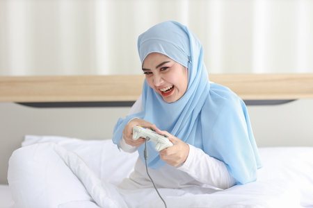VDO game console station concept. active asian woman wearing white muslim sleepwear sitting on bed, holding joystick and playing exciting game. Cute girl looked excited with game controller consoleの写真素材