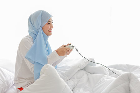VDO game console station concept. active asian woman wearing white muslim sleepwear sitting on bed, holding joystick and playing exciting game. Cute girl looked excited with game controller consoleの写真素材