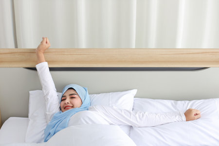Beautiful asian woman wearing white muslim sleepwear lying on bed, stretching her arms after getting up in the morning at sunrise. Cute young girl with hijab wake up and relaxing, closing her eyesの写真素材