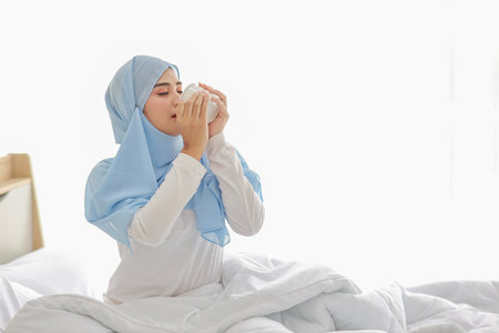 Beautiful and healthy asian woman sitting on bed and drinking coffee or tea after getting up in the morning at sunrise in bedroom with happy and smiling face.の写真素材