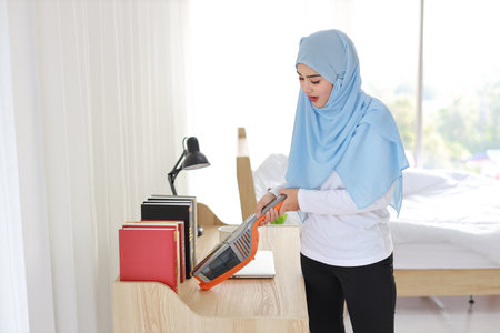 Closeup active young asian muslim housewife woman hands cleaning with vacuum cleaning wooden table with computer blurred background. Cleaning girl in white dress and black leggings vacuuming her roomの写真素材