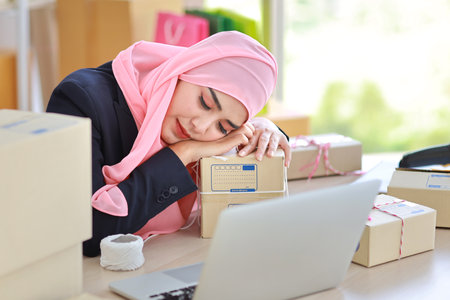 Religious asian woman in blue muslim suit and pink shaft frustrated laid her head down on the table, sit and work on wooden table with computer and package box delivery. Young girl sleep while workingの写真素材