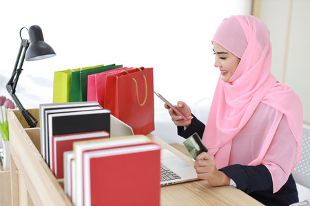 Active smart asian woman in blue muslim suit and pink shaft sitting and holding credit card while using mobile phone for payment or online purchasing. Startup small business girl work at home.の写真素材