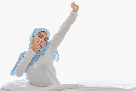 Beautiful asian woman stretching her arms after getting up in the morning on bed at sunrise in bedroom. Cute young girl in muslim dress and hijab sitting and relaxing with happy and smiling face.の写真素材
