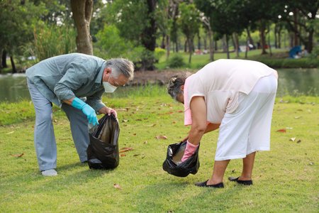volunteer tourist asian senior couple cleaning up garbage and plastic debris on dirty park by collecting them into big black bagの写真素材