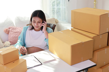 Attractive happy asian pregnant woman sitting and using mobile smart phone and writing order for customer with tablet. Expectant mother preparing and working for baby birth during pregnancy concept.の写真素材