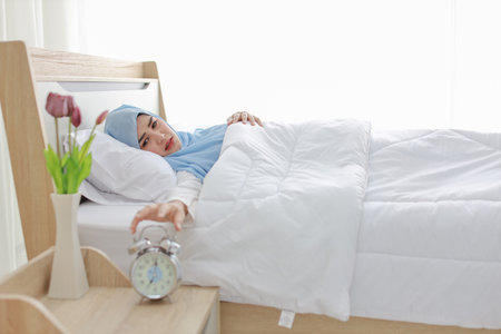 Oversleeping asian woman white muslim wearing sleepwear lying on bed, missing ring of alarm clock waking up. Portrait young girl trying to stop ringing in the morningの写真素材