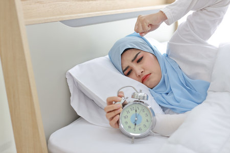 Oversleeping asian woman white muslim wearing sleepwear lying on bed, missing ring of alarm clock waking up. Portrait young girl trying to stop ringing in the morningの写真素材