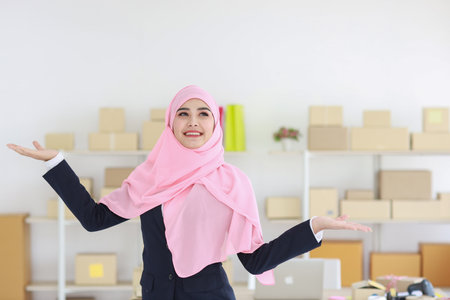 Young religious asian woman in blue muslim suit and pink shaft on head standing with confidence showing something. Business woman stand with package SME box delivery background. Work at home conceptの写真素材