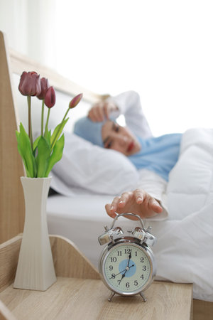 Oversleeping asian woman wearing white muslim sleepwear lying on bed, missing ring of alarm clock waking up. Side view portrait young girl hand trying to stop ringing while her hand on her faceの写真素材