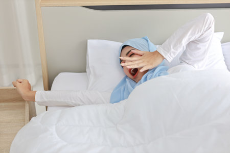 Beautiful asian woman wearing white muslim sleepwear lying on bed, stretching her arms after getting up in the morning at sunrise. Cute young girl with hijab wake up and relaxing, closing her eyesの写真素材