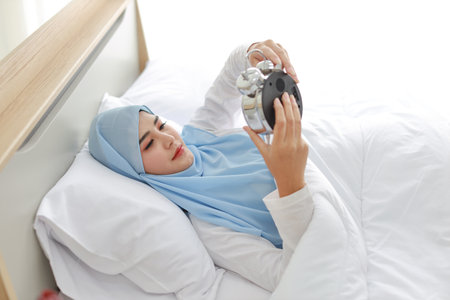Oversleeping asian woman wearing white muslim sleepwear lying on bed, missing ring of alarm clock waking up. Portrait young girl trying to stop ringing in the morningの写真素材