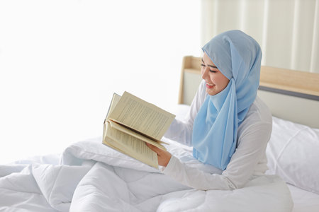 Happy young beautiful asian woman in muslim sleepwear sitting, reading book on bed. Cheerful and cute girl resting in bedroom and copy space for your advertising content.の写真素材