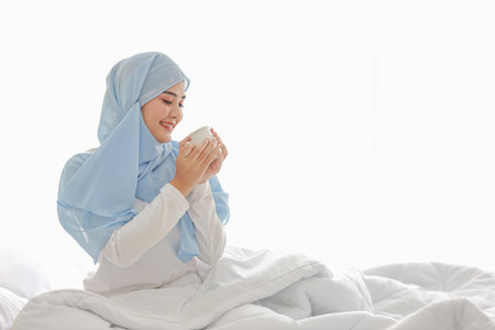 Beautiful and healthy asian woman sitting on bed and drinking coffee or tea after getting up in the morning at sunrise in bedroom with happy and smiling face.の写真素材